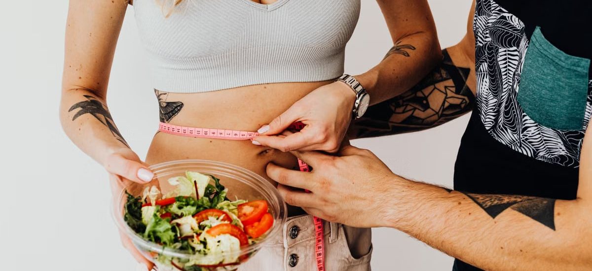 A couple measuring waistline with a tape and holding a fresh salad bowl, focusing on fitness and weight loss.