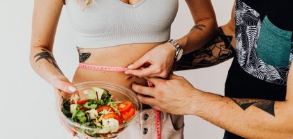 A couple measuring waistline with a tape and holding a fresh salad bowl, focusing on fitness and weight loss.