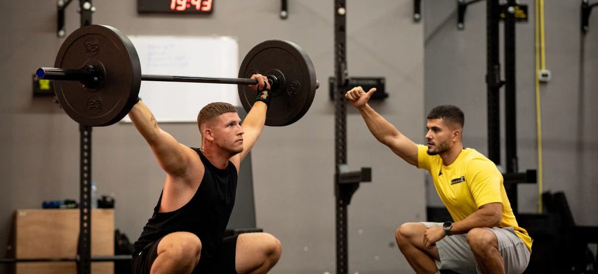 Man lifting weights under coach supervision in a gym setting, focusing on strength training.