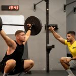 Man lifting weights under coach supervision in a gym setting, focusing on strength training.